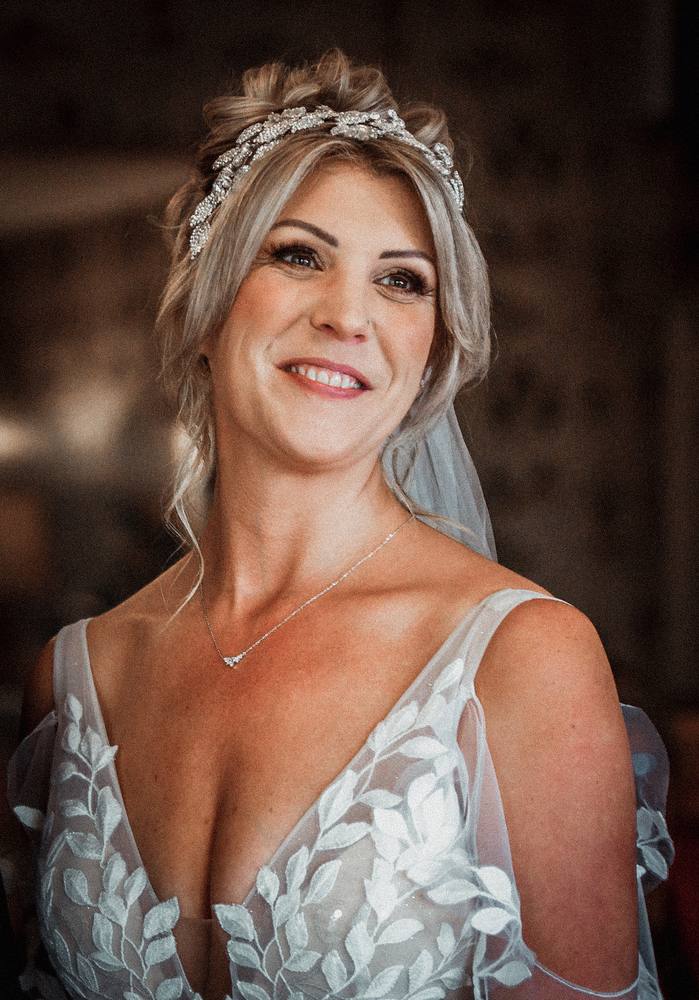 A smiling bride in a jewelled leaf headpiece and lace gown, looking off to one side
