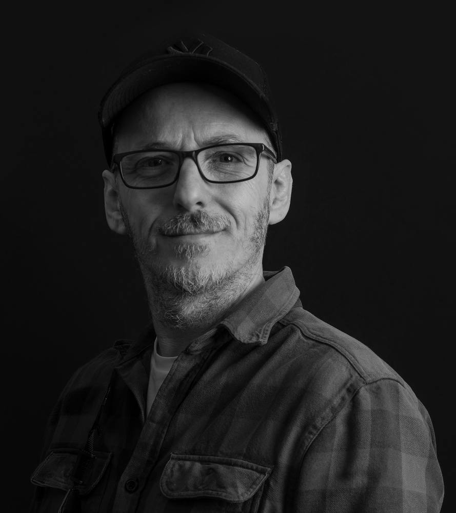 Black and white portrait of photographer Mark Kaye, smiling, in a cap and glasses