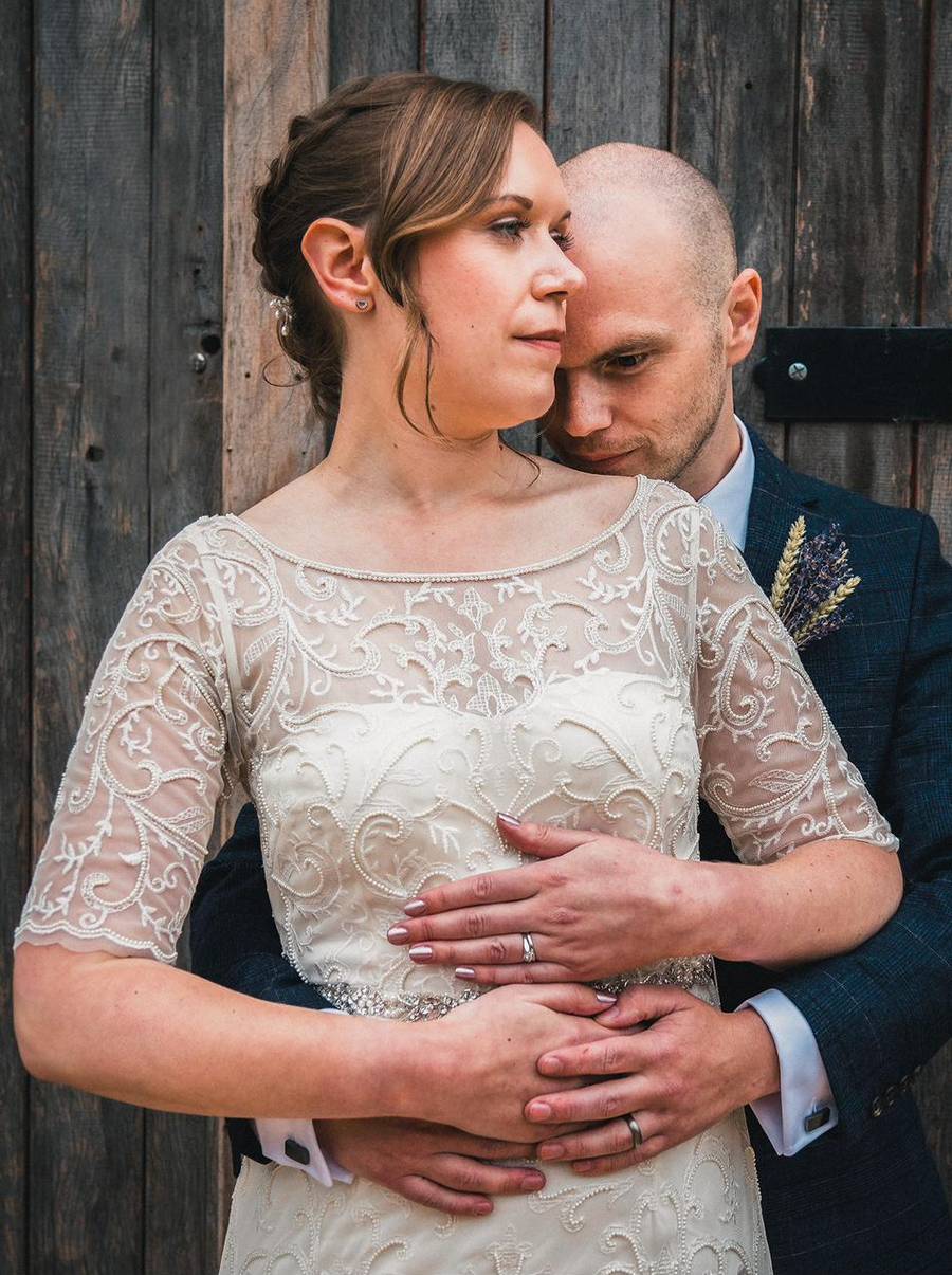 A groom holding his bride from behind against a weathered wooden door, her lace dress catching the light