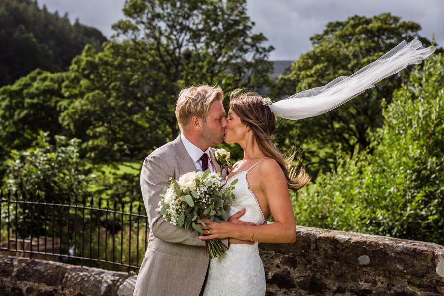 Bride and groom kissing beside a stone wall as her veil lifts in the wind